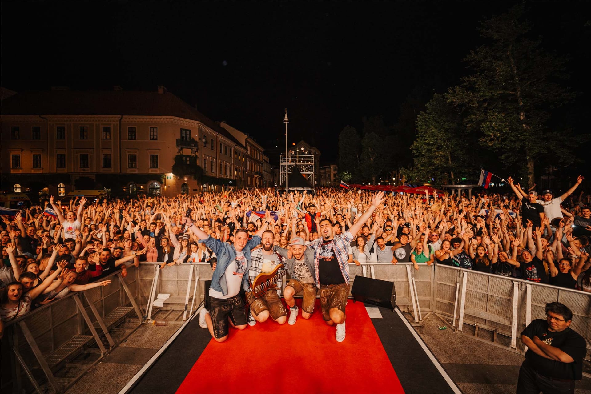 Nastopajoči pozirajo pred množico na odru z rdečo preprogo, večerni koncert.