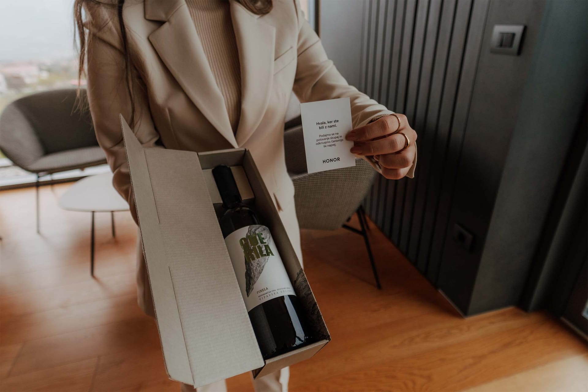 Ženska v svetlem plašču drži darilno škatlo s steklenico vina in voščilnico.