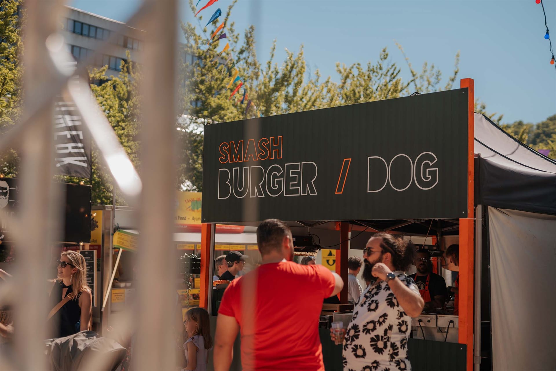 Ljudje pred stojnico s hrano z napisom Smash Burger / Dog na ulični tržnici.